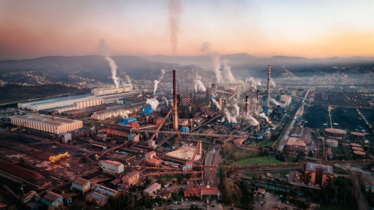 Blog Aerial view of a factory with chimneys emitting smoke during sunset, highlighting industrial pollution.