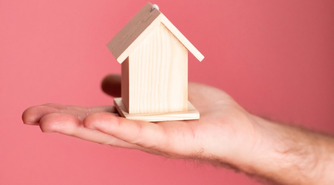 Wooden house model in hand symbolizing real estate concepts on pink background.