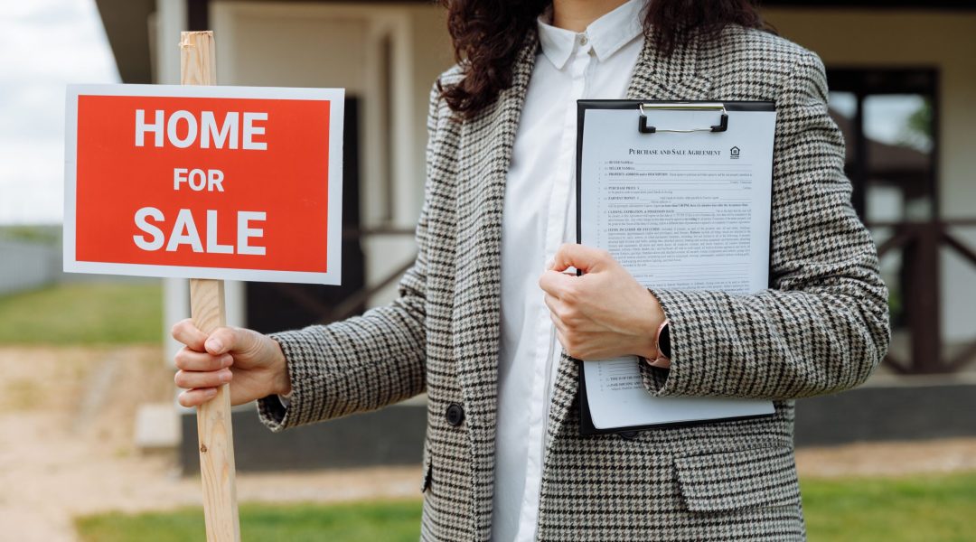 A real estate agent holding a home for sale sign and clipboard outside a property.